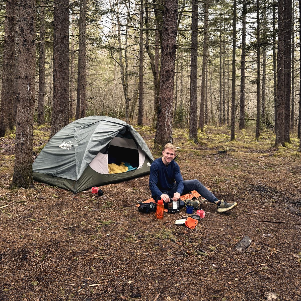 Alex at camp in an old spruce stand, breakfast going on the stove outside the tent.