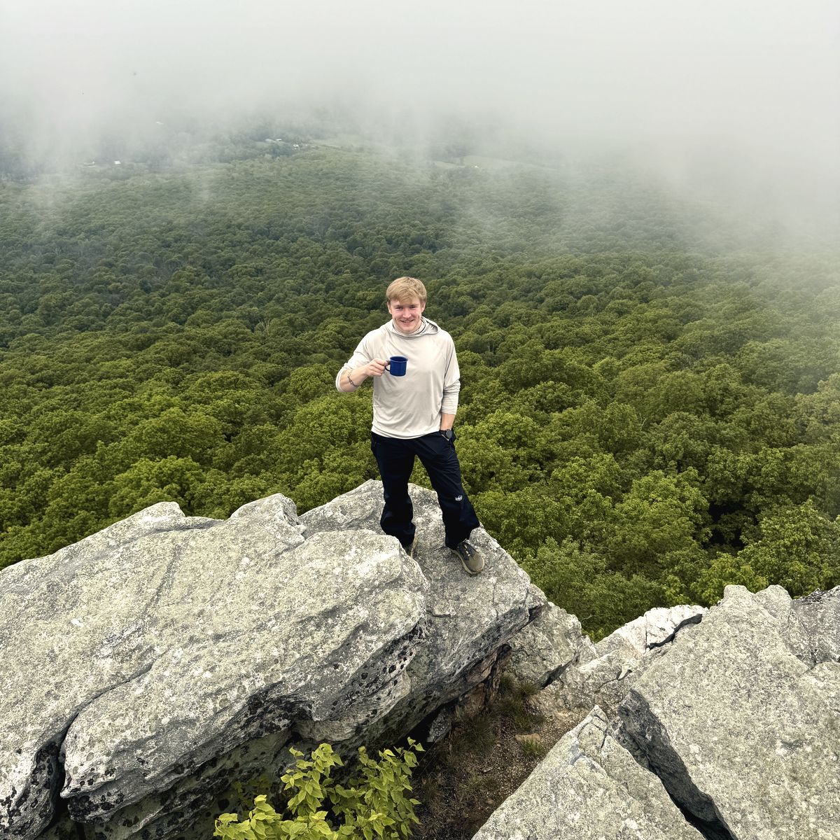 Alex on a ridgeline outcrop with a blue enamel mug, cloud sitting on the summit above the treeline.